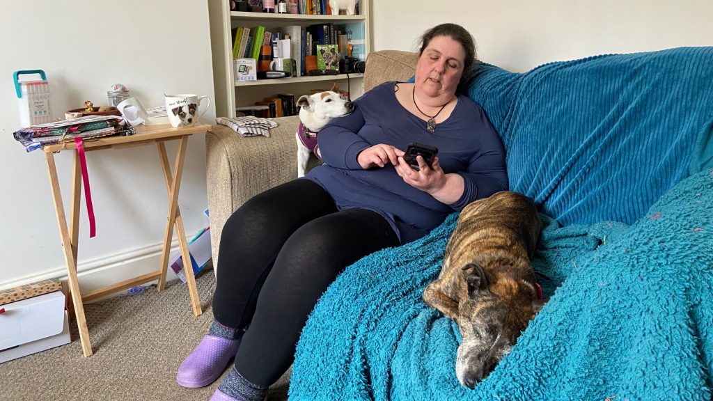 Burt, Jan and Muppet sitting on the couch with a bookshelf in the background. Jan is holdinga mobile phone creating Burt's Youtube channel. Burt is supervising while Muppet sleeps