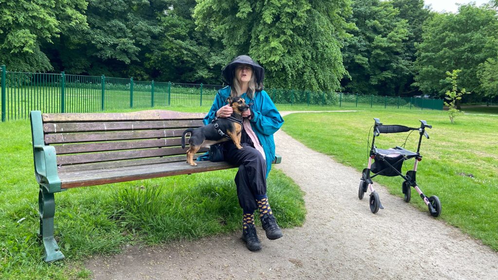 Madeleine and Cuthbert on a bench at Hale Park with the walker at the side on the path
